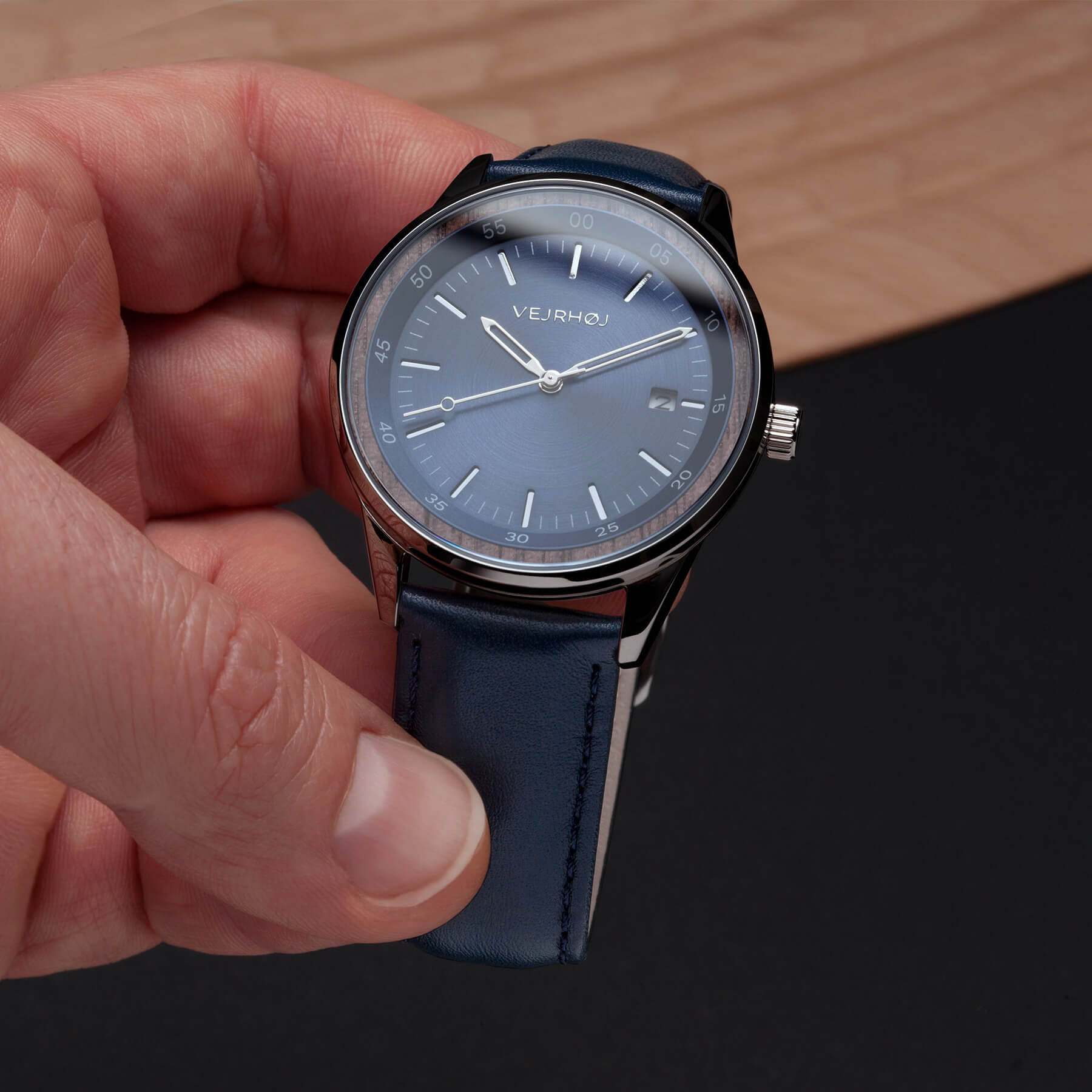 手が文字盤の青く、針のシルバー色の機械式腕時計を持って見せている。文字盤が木の輪で囲まれている