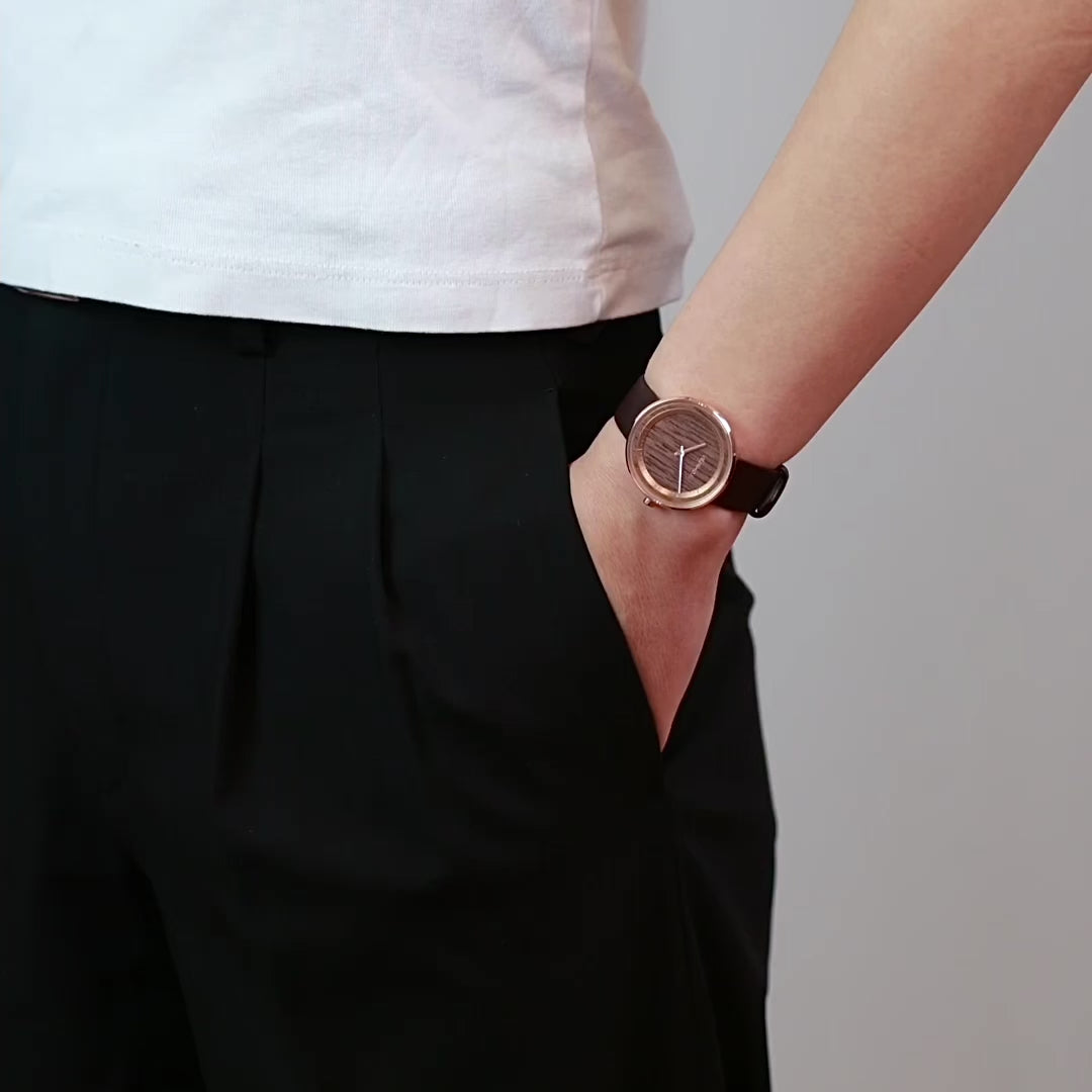 ローズゴールドメタルの腕時計を手首に付けている人が手をポケットの中に入れたままが立っている