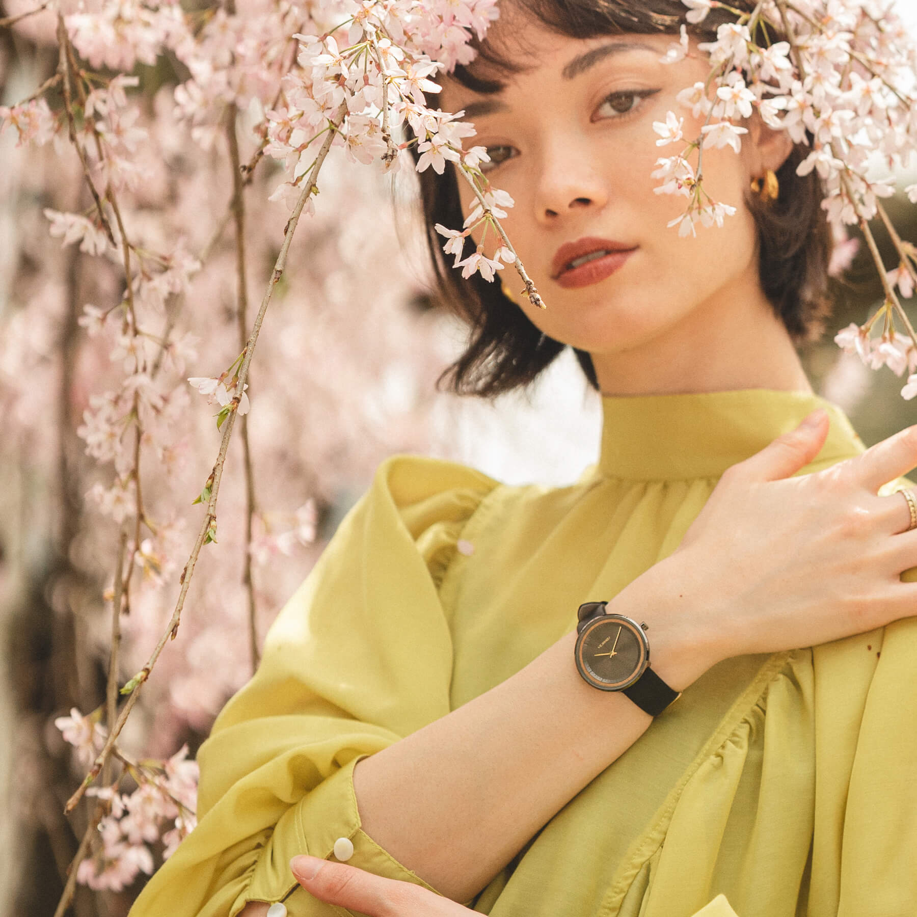 ヴェアホイの黒の腕時計を身に付けながら桜の木の後ろでポーズをとる女性